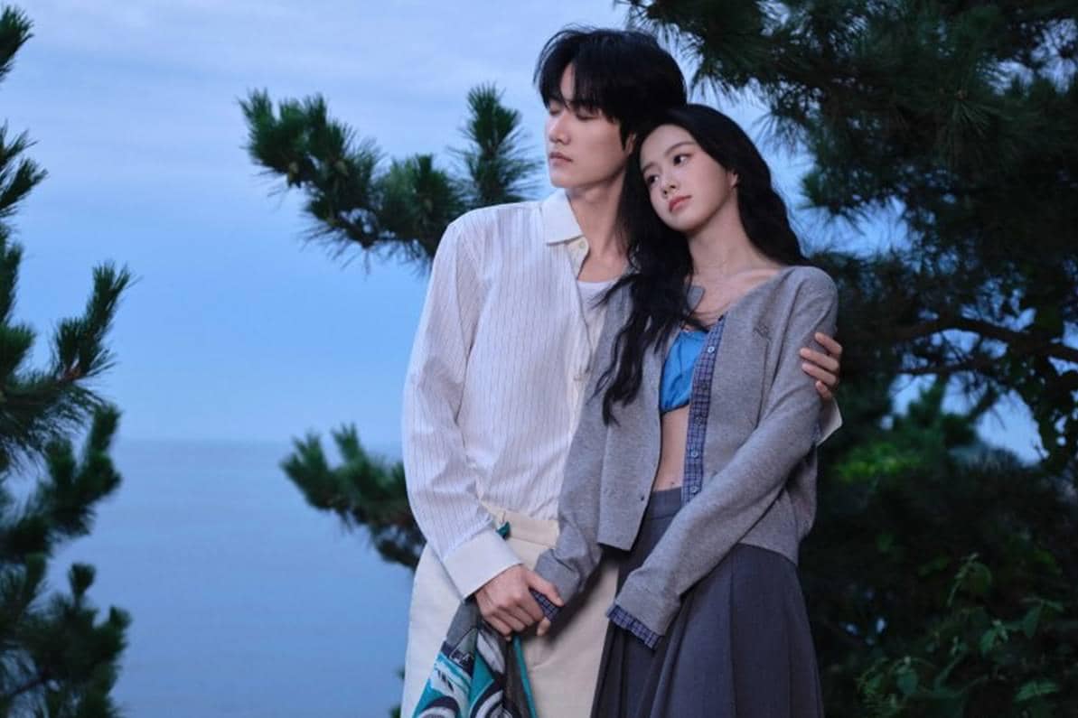 Sepasang pria dan wanita berdiri berdekatan di tepi laut saat senja, dikelilingi pepohonan hijau dengan suasana romantis dan tenang.