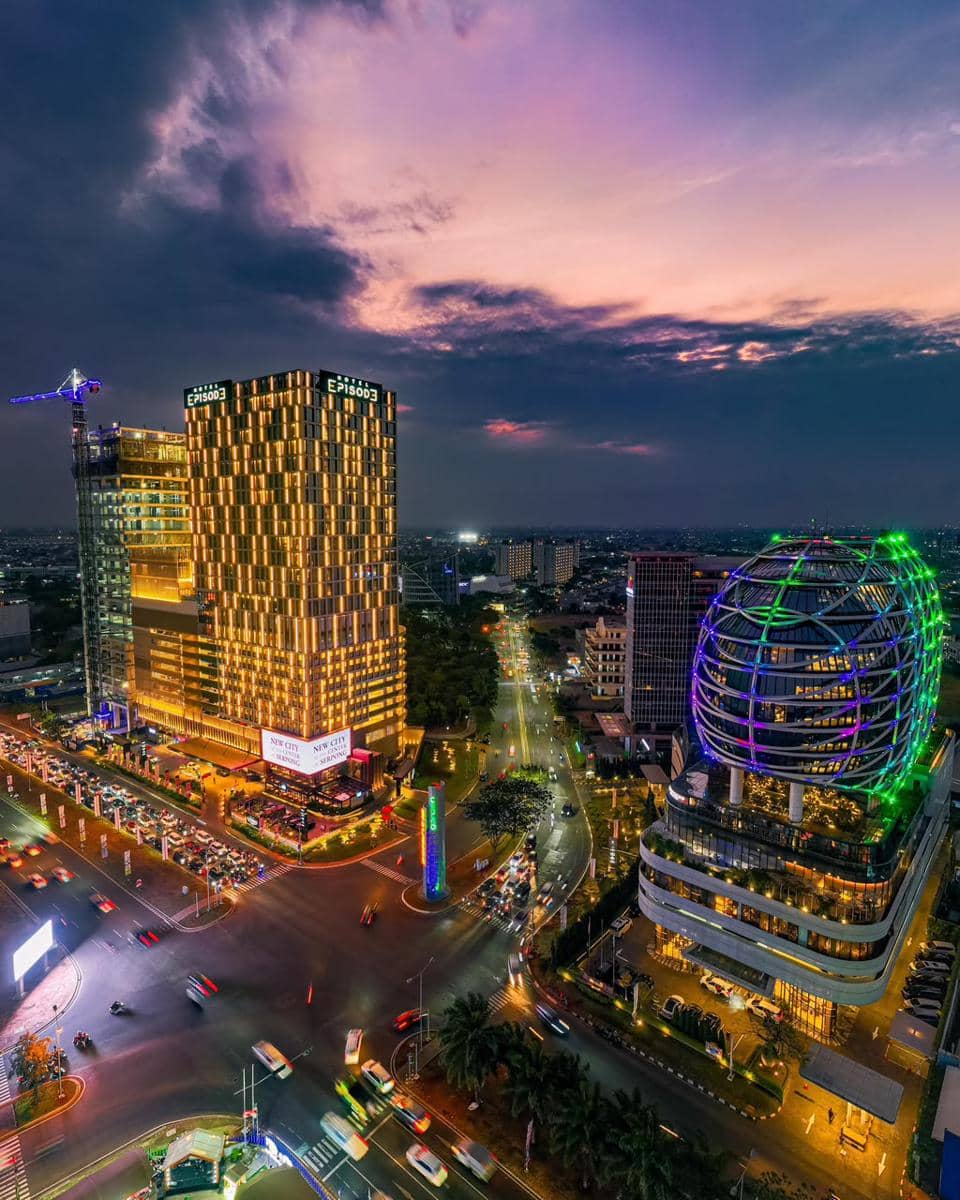 Pemandangan malam kawasan Gading Serpong dengan gedung tinggi bercahaya dan lalu lintas ramai di bawah langit senja berwarna ungu.