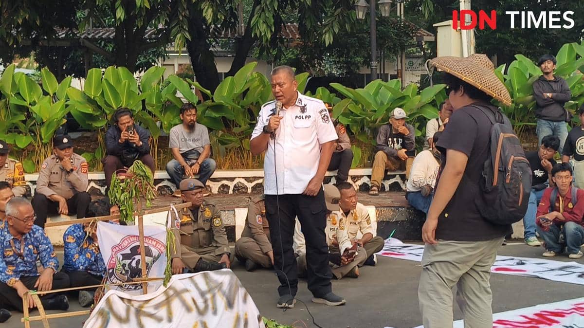 Kepala Satpol PP Kabupaten Bogor berbicara menggunakan mikrofon di hadapan massa aksi yang duduk di jalan dengan latar pepohonan hijau.