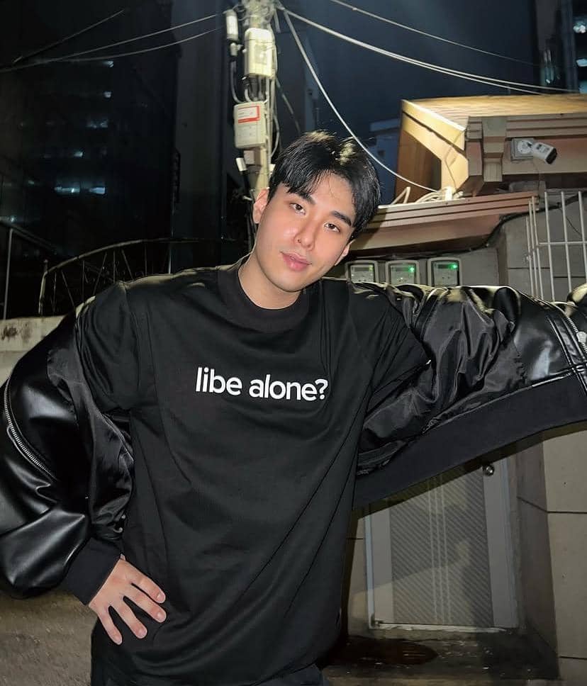 Seorang pria berpose santai di malam hari mengenakan kaus hitam bertuliskan 'libe alone?' dengan latar bangunan dan kabel listrik.