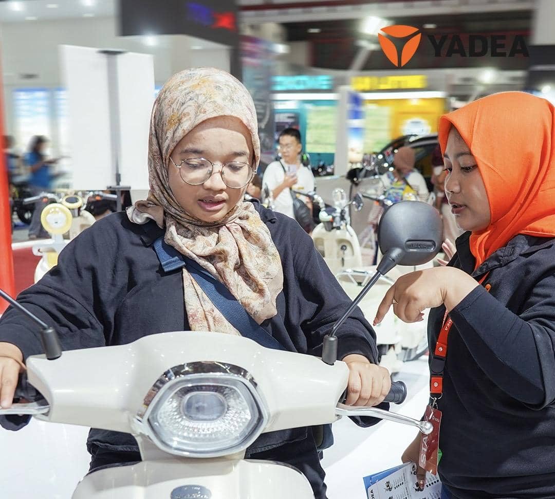 Dua perempuan di booth Yadea PRJ 2026, salah satunya mencoba motor listrik sambil berbincang dengan staf berpakaian hitam dan jilbab oranye.