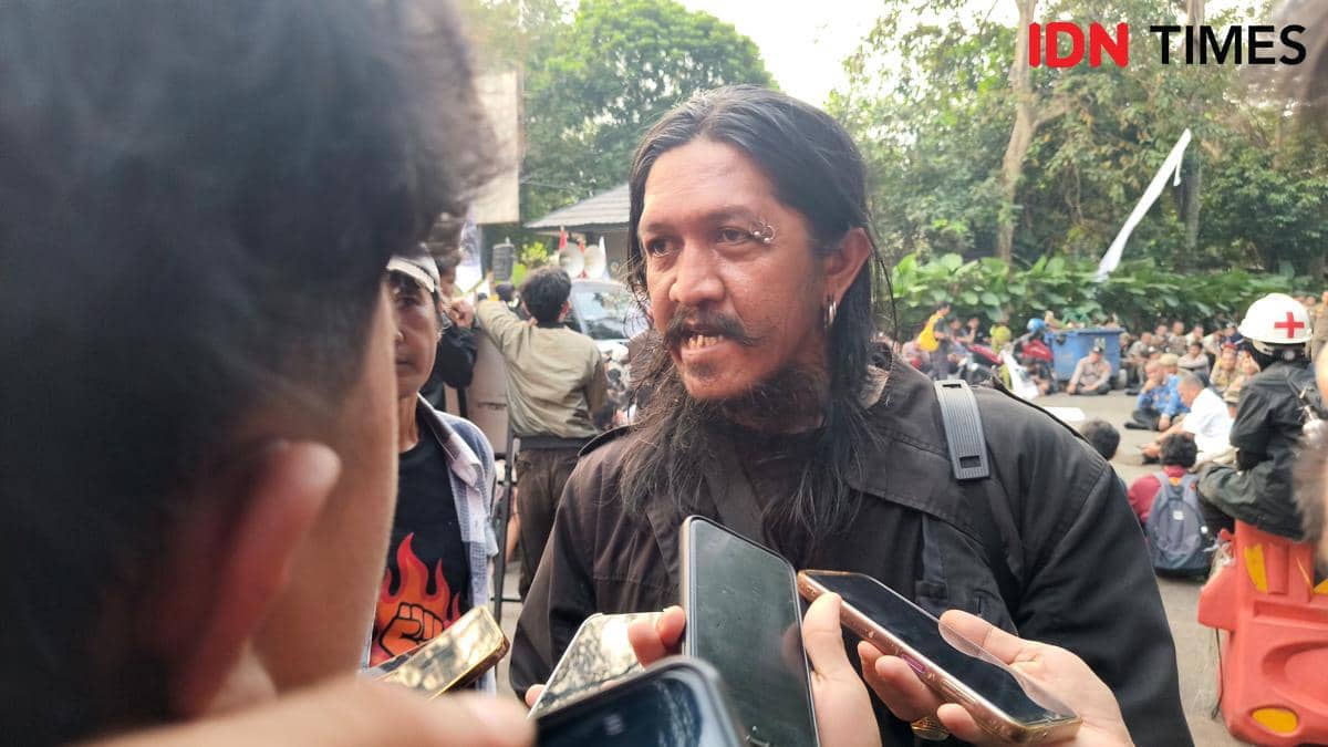 Seorang pria berambut panjang berbicara kepada wartawan yang memegang ponsel dan alat perekam di tengah kerumunan massa di luar ruangan.