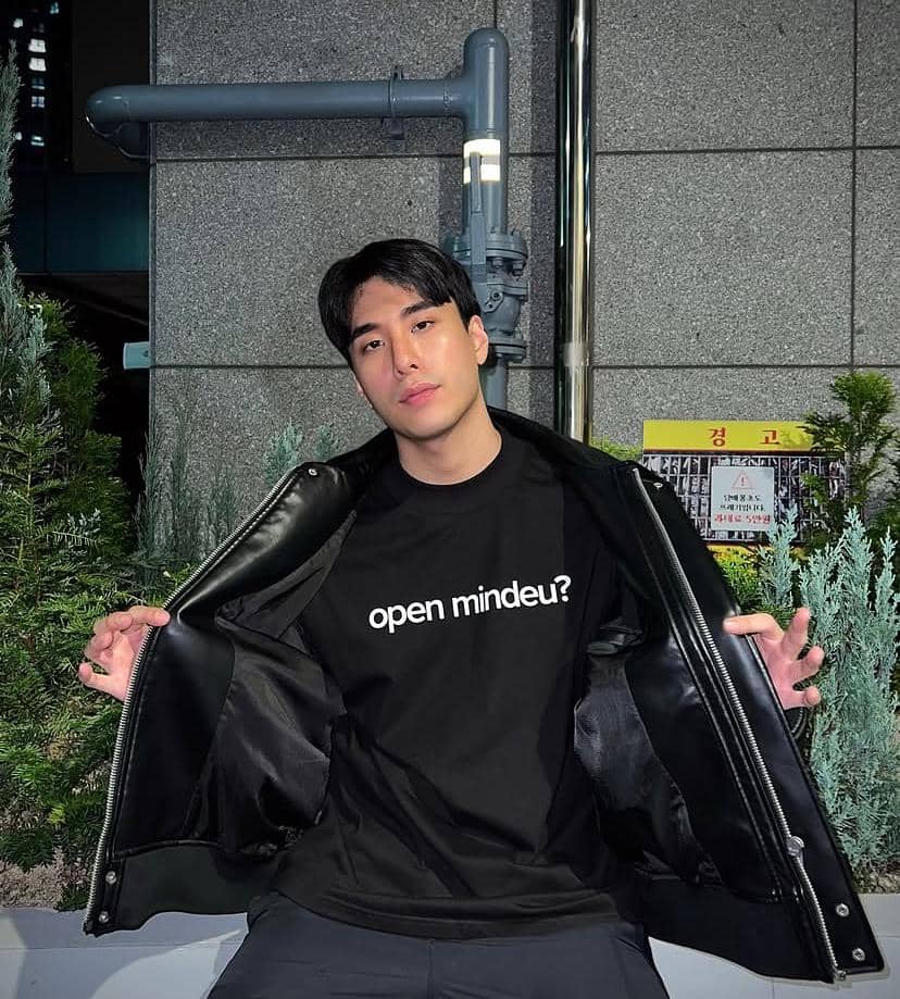Seorang pria berpose di luar ruangan mengenakan jaket hitam terbuka dan kaus hitam bertuliskan 'open mindeu?' dengan latar dinding abu-abu dan tanaman hijau.