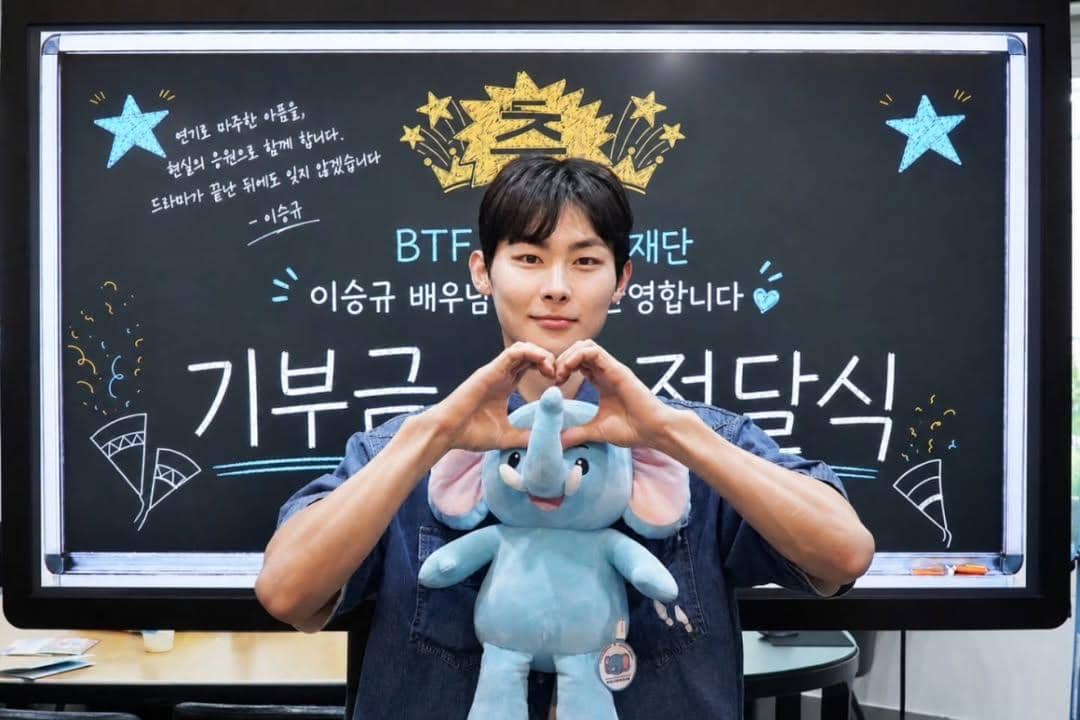 Seorang pria berpose membentuk hati dengan tangan sambil memegang boneka gajah biru di depan papan tulis bertema donasi.