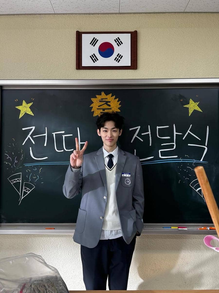 Seorang siswa mengenakan seragam sekolah abu-abu berpose dengan tanda damai di depan papan tulis bertuliskan huruf Korea dan bendera Korea Selatan di atasnya.
