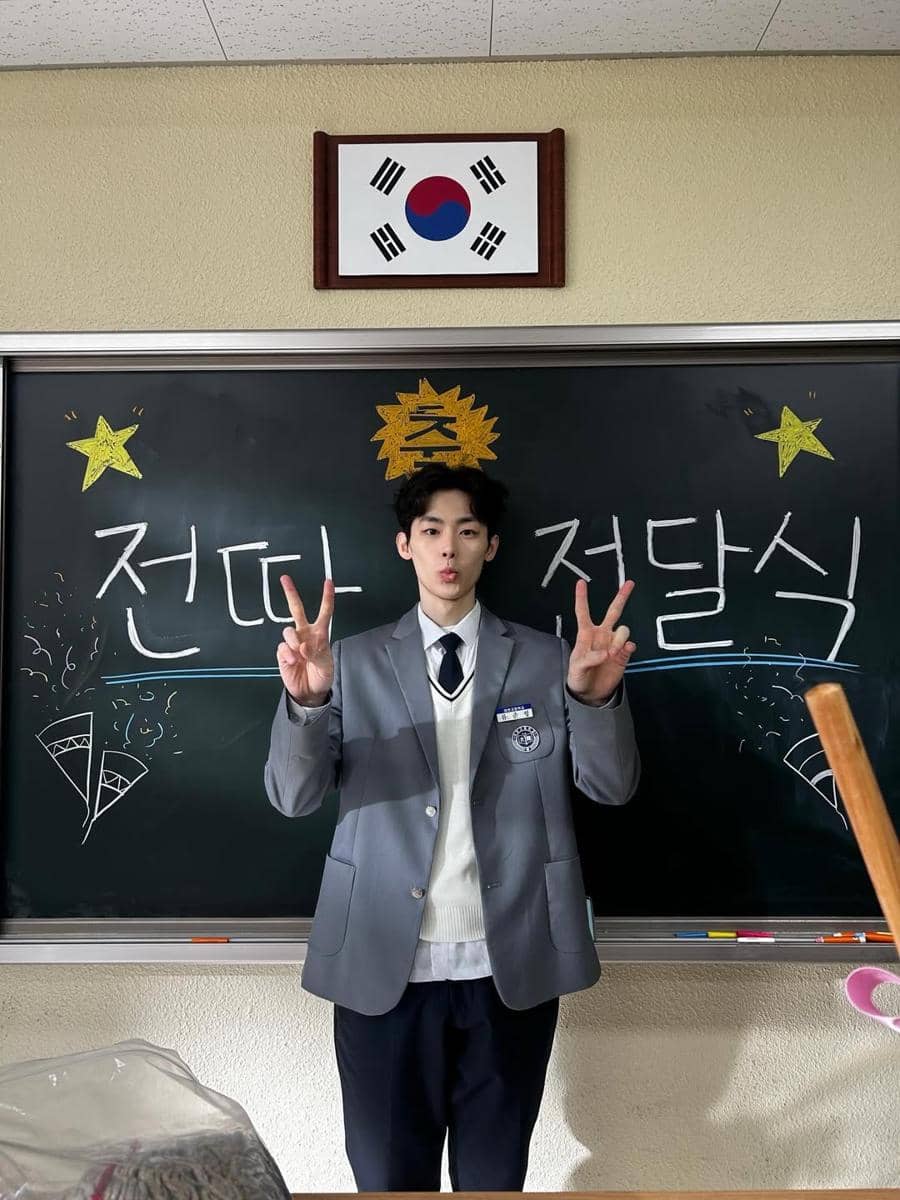 Seorang siswa berseragam sekolah abu-abu berpose dengan tanda damai di depan papan tulis bertuliskan huruf Korea dan bendera Korea Selatan di atasnya.