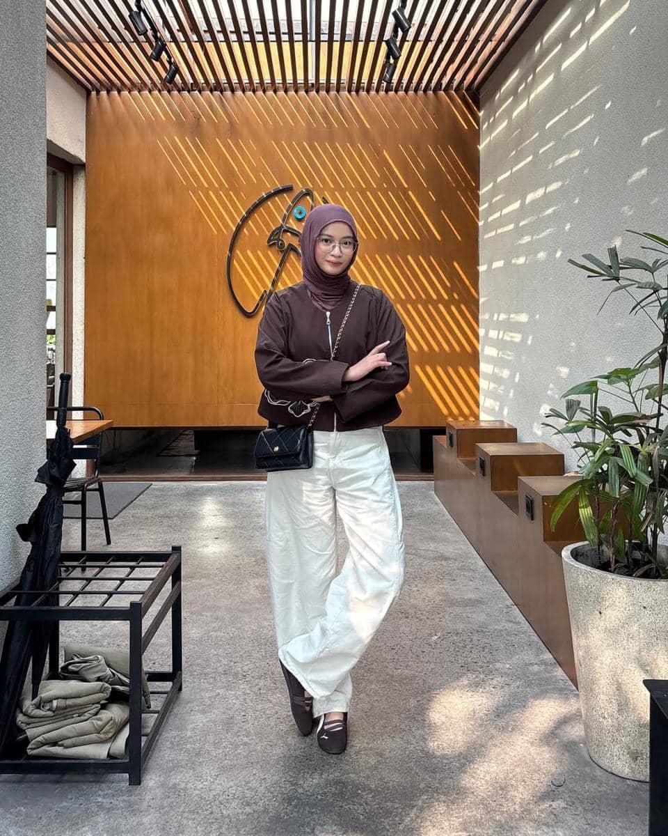 Seorang wanita berhijab mengenakan jaket cokelat, celana putih longgar, dan sneakers Puma Speedcat Ballet berwarna cokelat di area kafe modern.