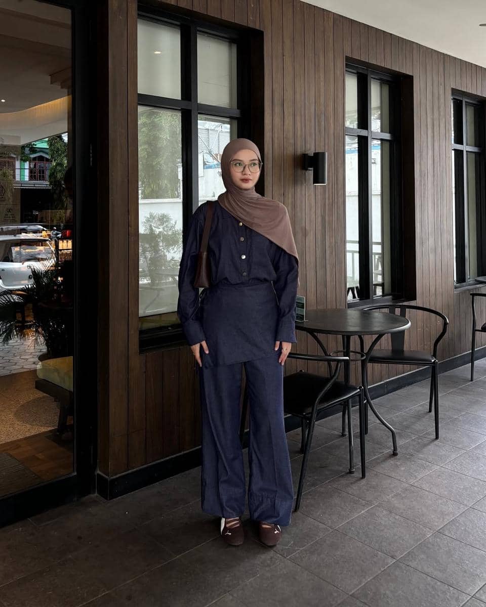 Seorang wanita berhijab mengenakan outfit bernuansa denim biru tua dengan sneakers Puma Speedcat Ballet berdiri di depan kafe berpanel kayu.