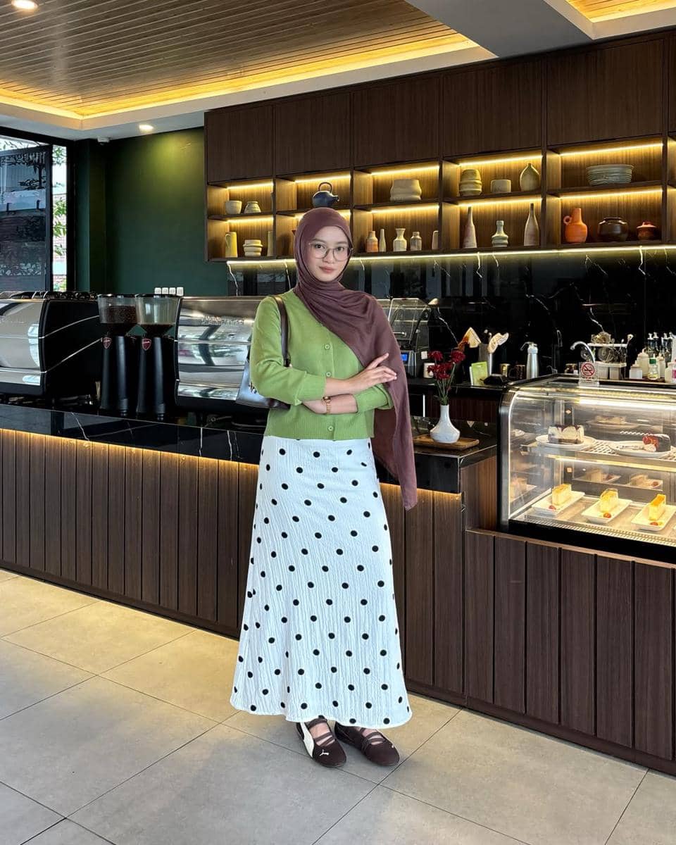 Seorang wanita berhijab mengenakan cardigan hijau, rok putih bermotif polkadot, dan sneakers Puma Speedcat Ballet berdiri di depan counter kafe modern.