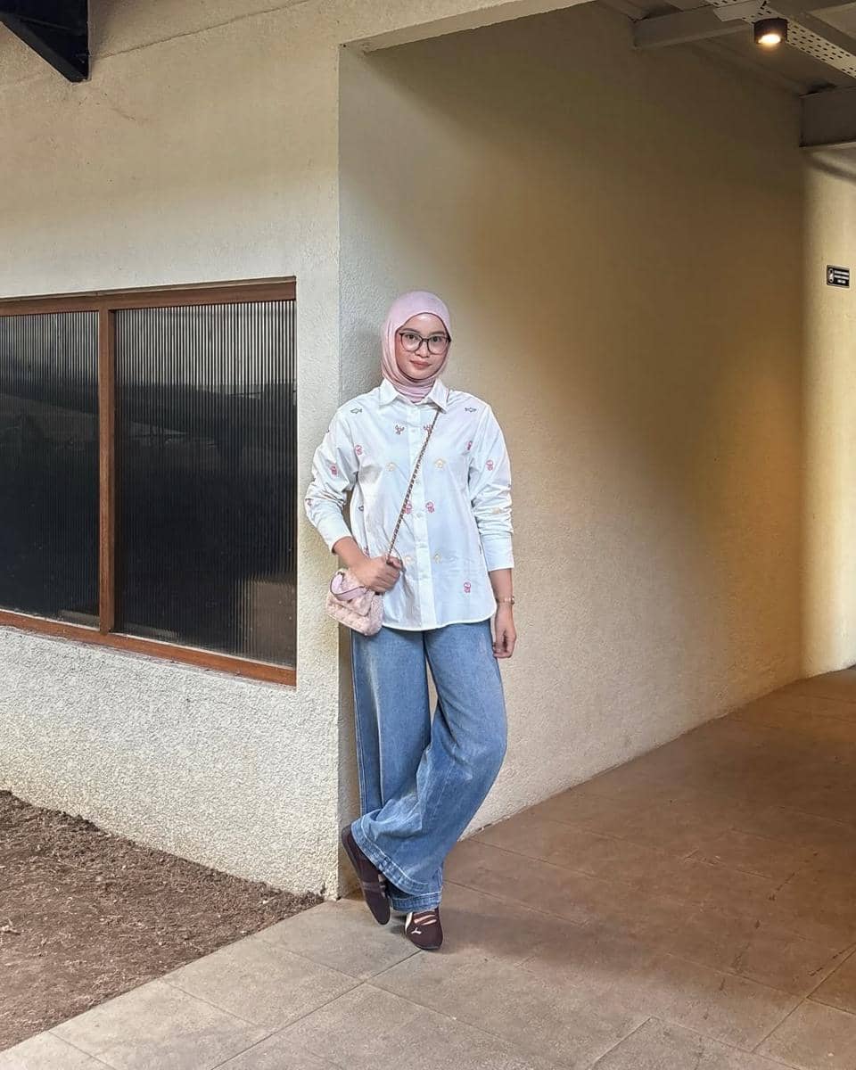 Seorang wanita berhijab mengenakan kemeja putih bermotif, celana jeans longgar, sneakers Puma Speedcat Ballet, dan tas kecil berwarna muda.