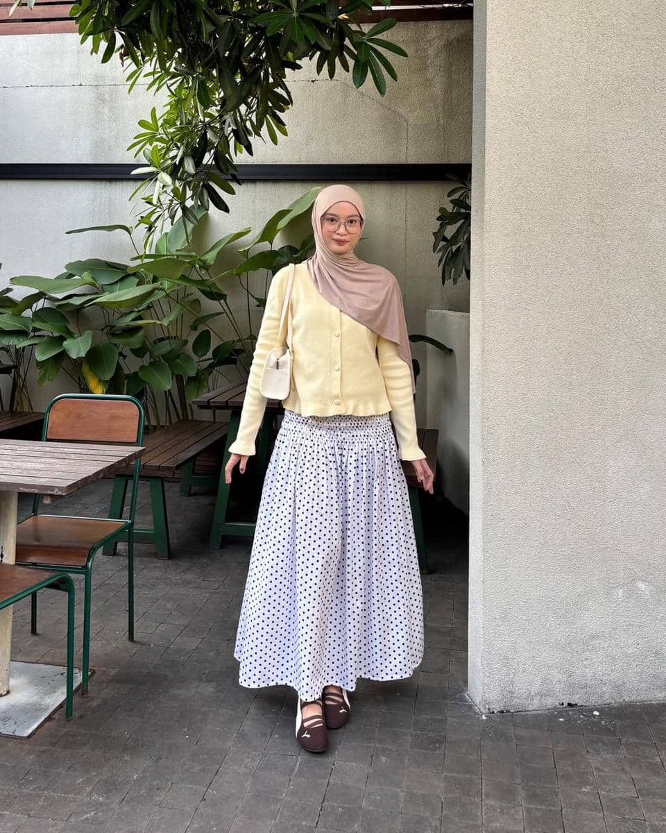 Seorang wanita berhijab mengenakan cardigan krem, rok panjang motif polkadot, dan sneakers cokelat bergaya kasual di area outdoor.