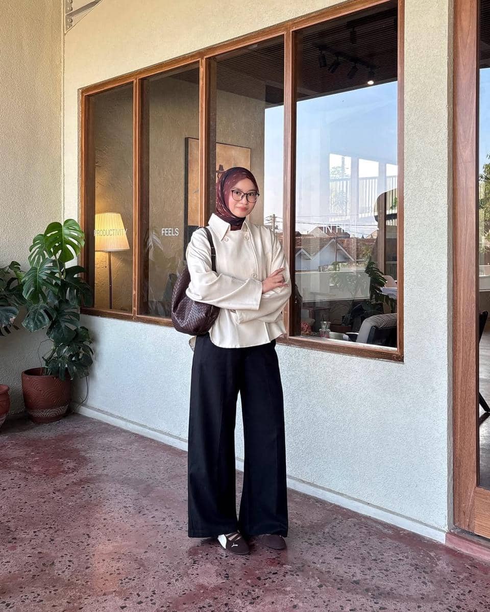 Seorang wanita berhijab mengenakan kemeja krem, celana hitam longgar, dan sneakers Puma Speedcat Ballet berwarna cokelat di depan jendela besar.