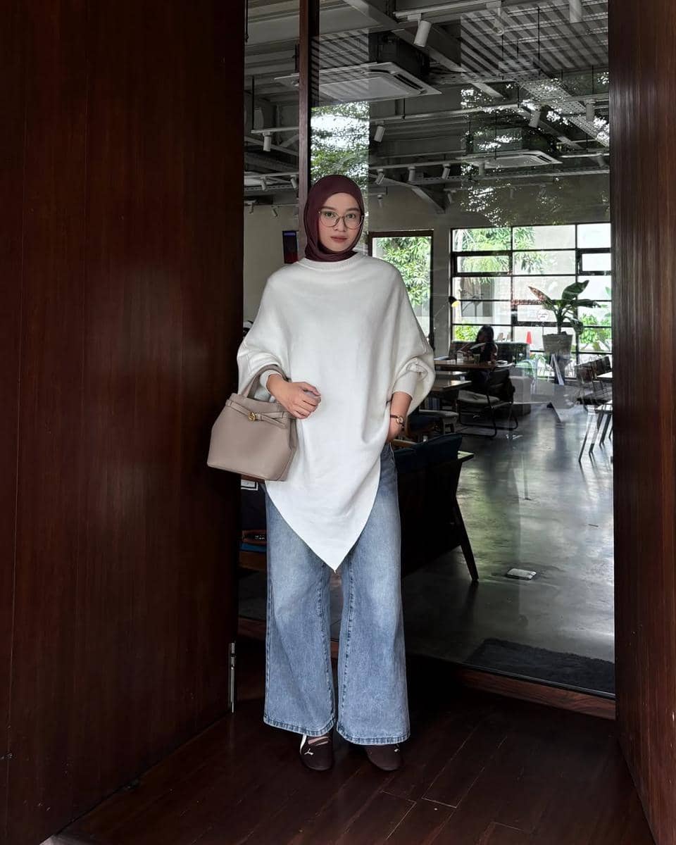 Seorang wanita berhijab mengenakan sweater putih longgar, celana jeans lebar, sneakers cokelat, dan tas tangan krem di depan pintu kayu.