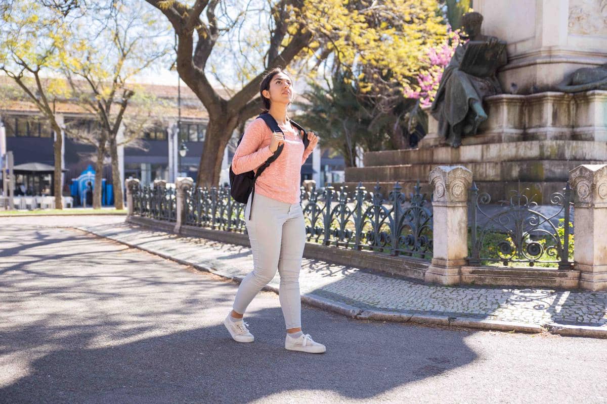 Seorang perempuan berjalan santai di taman kota dengan mengenakan tas ransel hitam dan pakaian kasual berwarna cerah di hari yang cerah.
