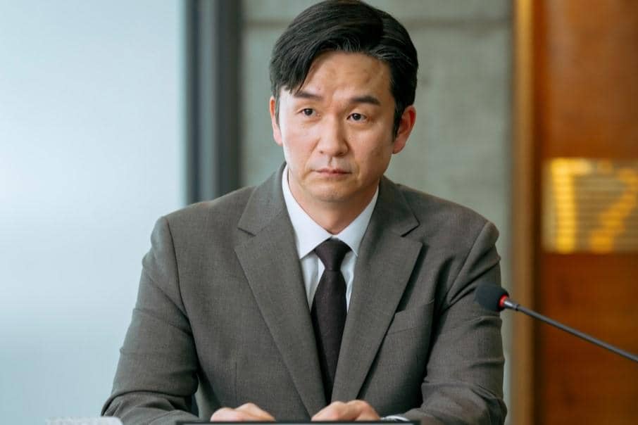 Seorang pria mengenakan setelan jas abu-abu duduk di meja dengan mikrofon di depannya dalam cuplikan drama Reborn Rookie.