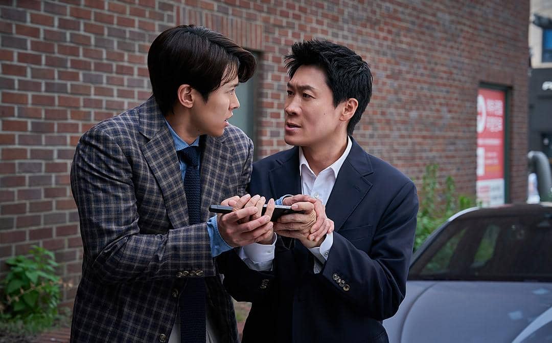 Dua pria berjas berdiri di depan dinding bata sedang berdebat sambil memegang ponsel dalam cuplikan drama Korea Husbands in Action.