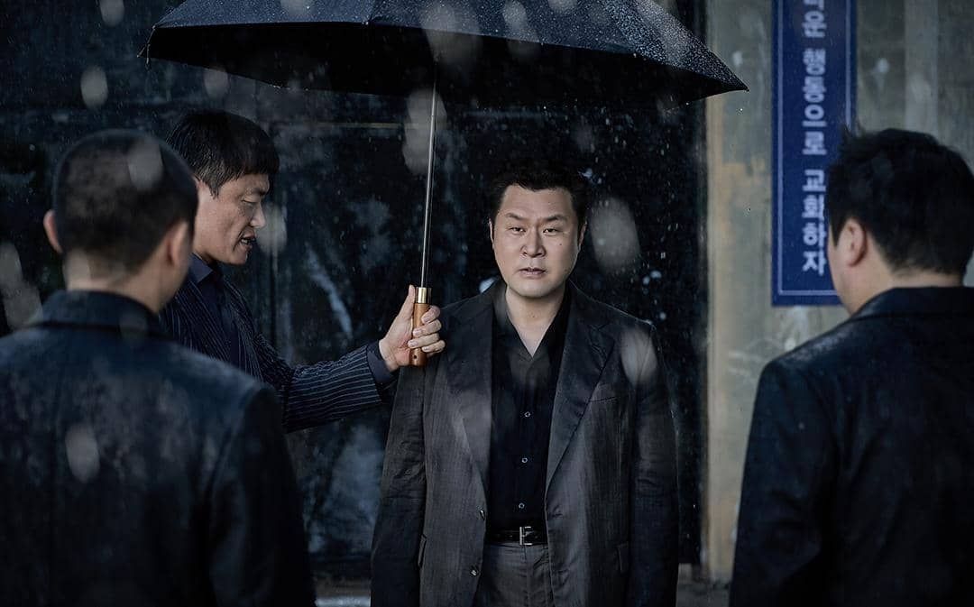 Beberapa pria berjas berdiri di bawah hujan, salah satunya memegang payung hitam dalam suasana tegang dari drama Korea Husbands in Action.