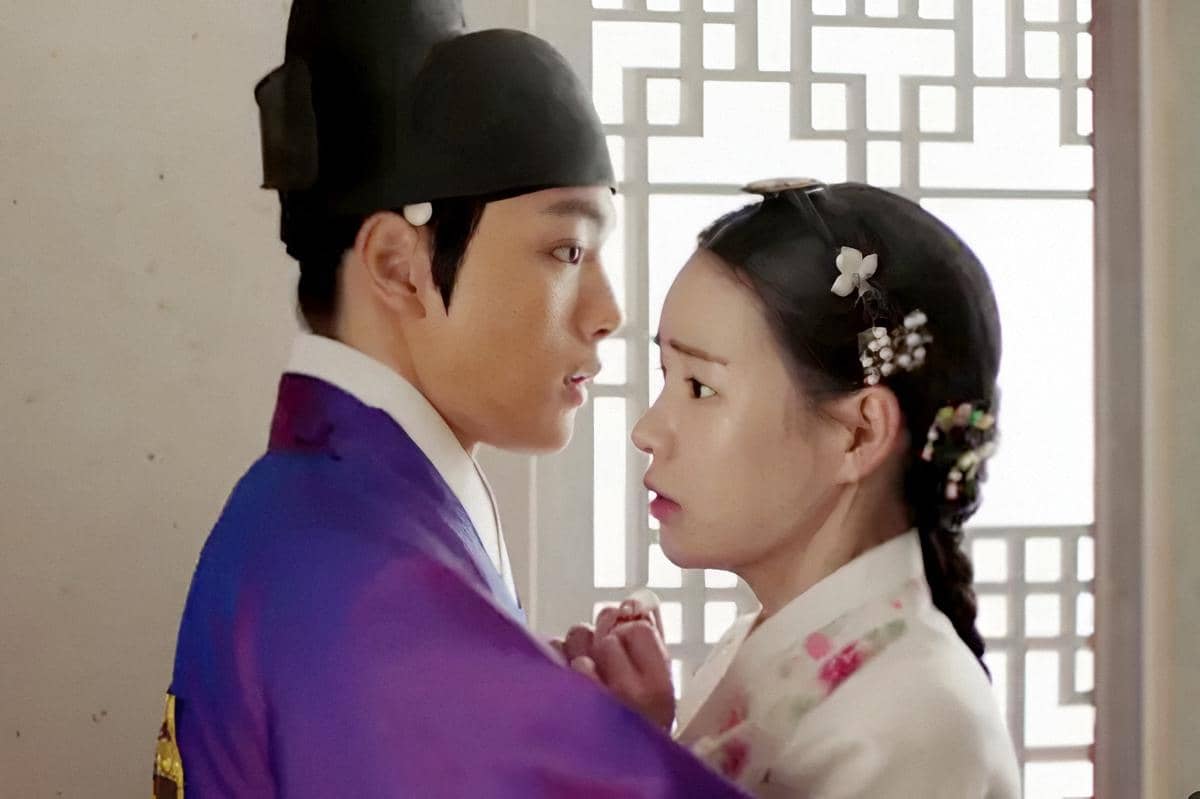 Dua karakter dalam drama The Royal Gambler mengenakan hanbok tradisional Korea, saling menatap dengan ekspresi tegang di dekat jendela berpola.