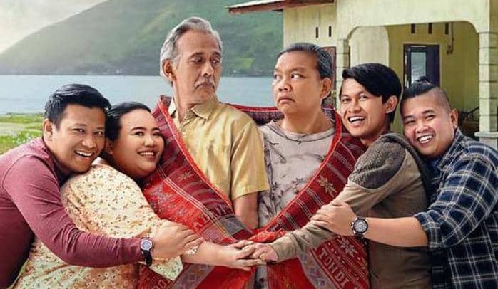 Enam orang berdiri berpelukan di depan rumah dengan latar pegunungan dan danau, dua di antaranya dibalut kain ulos merah.