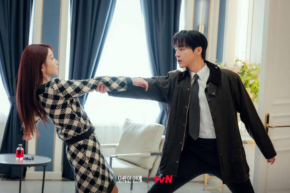 Dua karakter dalam drama Spooky in Love berdiri saling berhadapan di ruangan elegan, dengan salah satu menarik tangan yang lain.