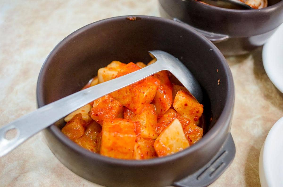 Potongan tofu berlumur saus gochujang merah disajikan dalam mangkuk hitam dengan sendok logam di atas meja.