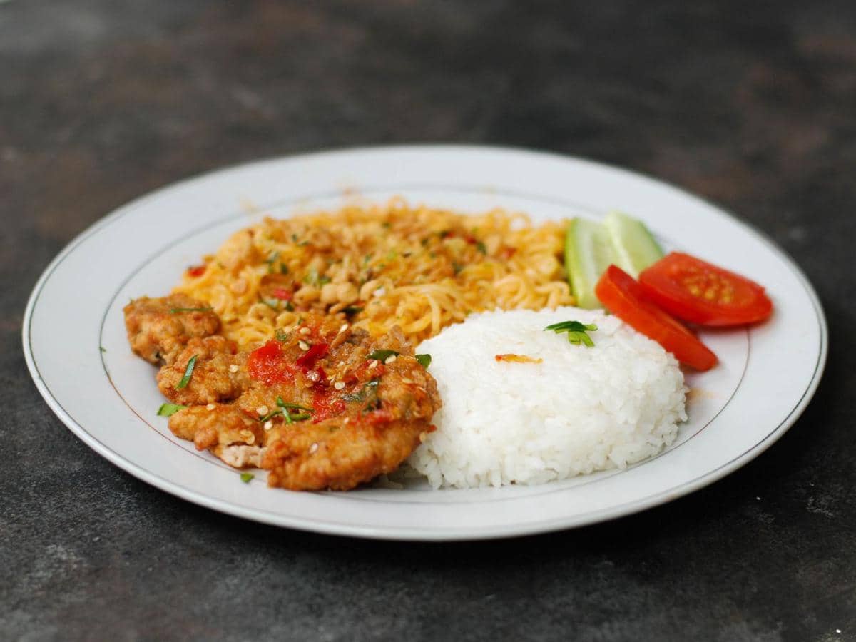 Sepiring tahu sambal geprek disajikan dengan nasi putih, mi goreng, serta irisan tomat dan mentimun di atas piring putih.