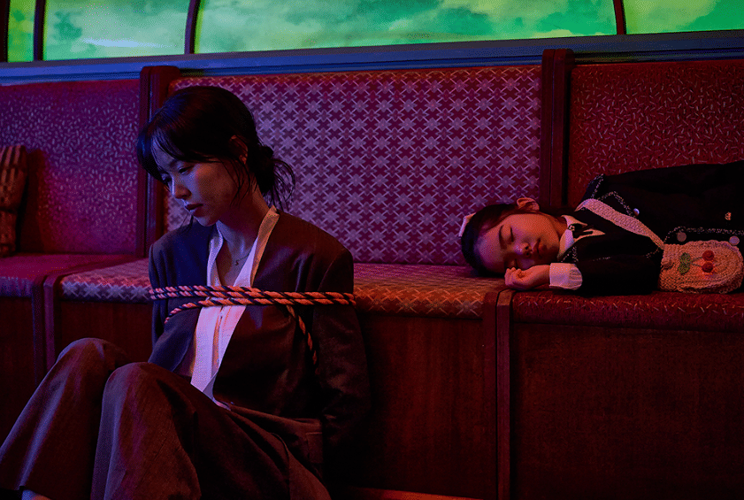 Seorang wanita terikat tali duduk di lantai sementara seorang anak perempuan tidur di bangku dalam adegan drama Korea Husbands in Action.