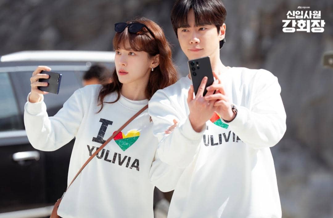 Dua orang mengenakan sweater putih bertuliskan 'I YULIVIA' sedang berfoto selfie di luar ruangan dengan latar mobil dan tebing.