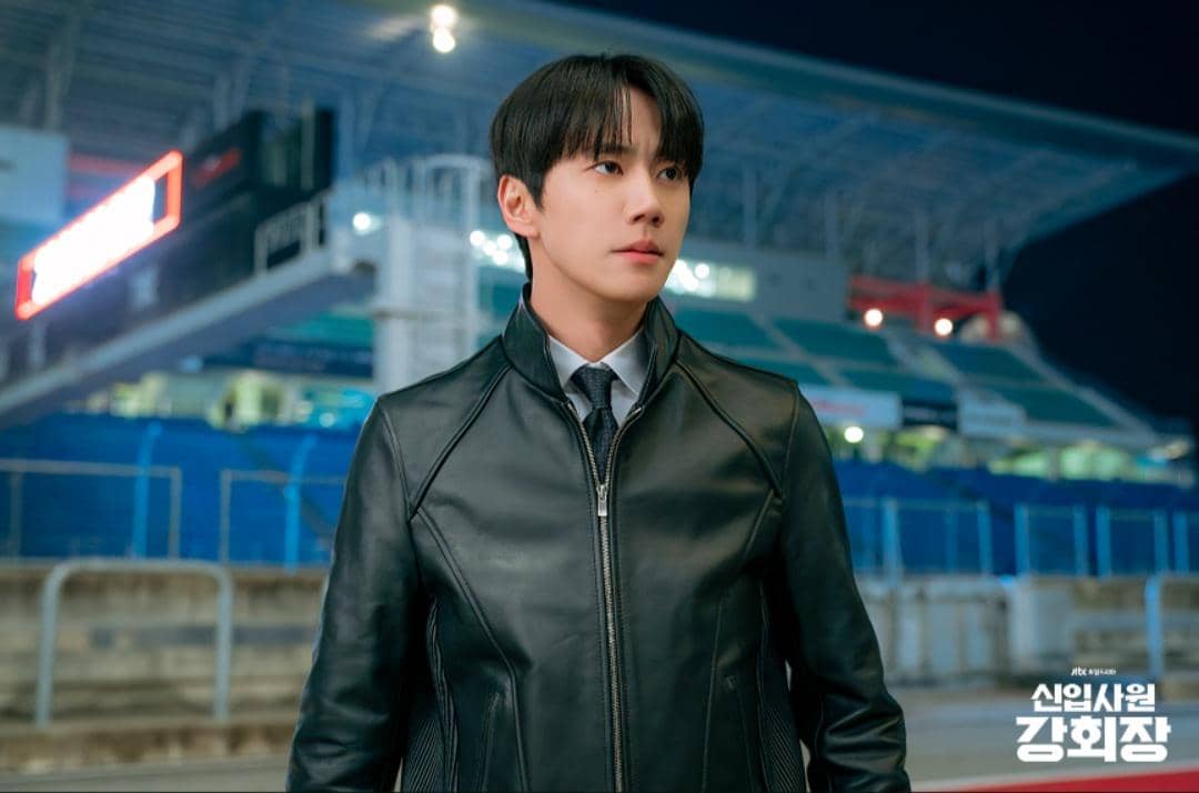 Seorang pria mengenakan jaket kulit hitam dan dasi berdiri di area stadion pada malam hari dalam cuplikan drama Reborn Rookie.