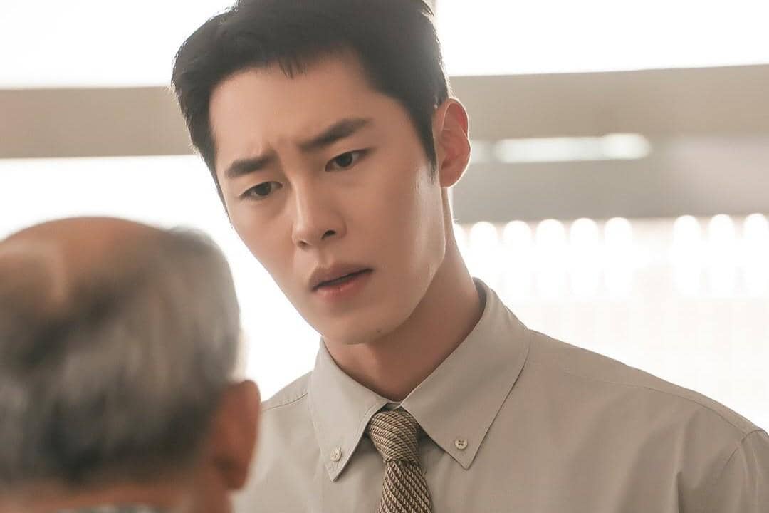 Seorang pria muda mengenakan kemeja abu-abu dan dasi sedang berbicara serius dengan pria tua di depannya dalam adegan drama Doctor on the Edge.