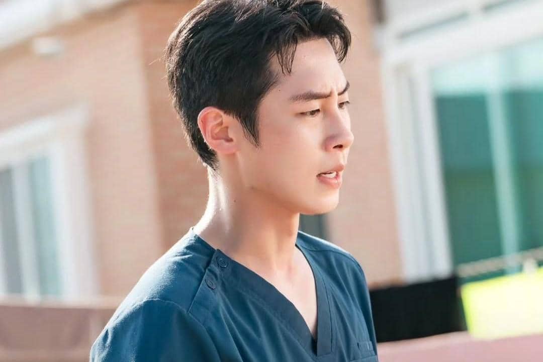 Seorang pria mengenakan seragam medis biru berdiri di luar ruangan dengan ekspresi serius dalam adegan drama Doctor on the Edge.