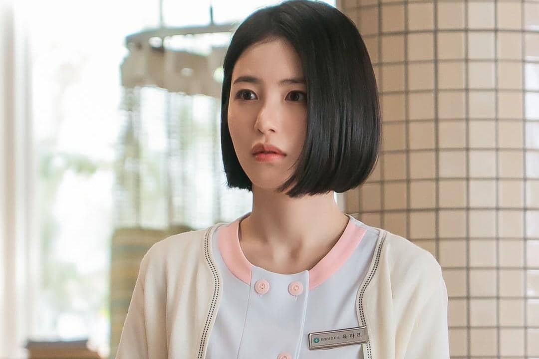 Seorang wanita berambut pendek mengenakan seragam medis berwarna putih dan pink dengan kartu nama di dada dalam adegan drama Doctor on the Edge.