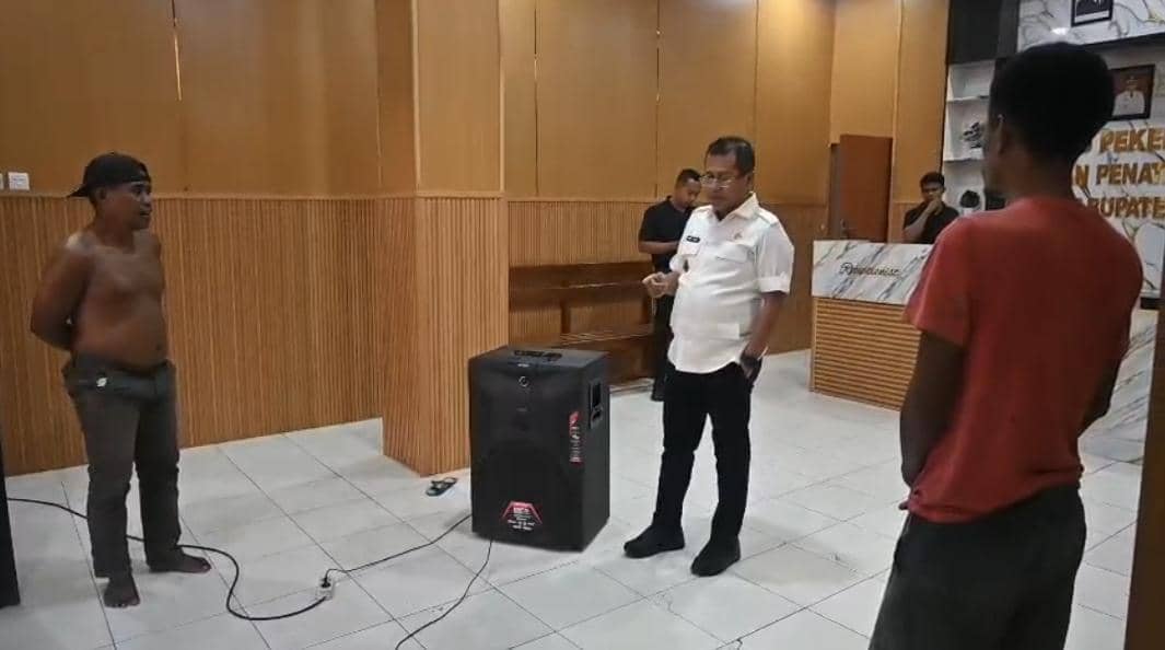 Beberapa pria berdiri di dalam ruangan kantor dengan dinding kayu, salah satunya mengenakan seragam putih dan berbicara kepada dua pria lainnya.
