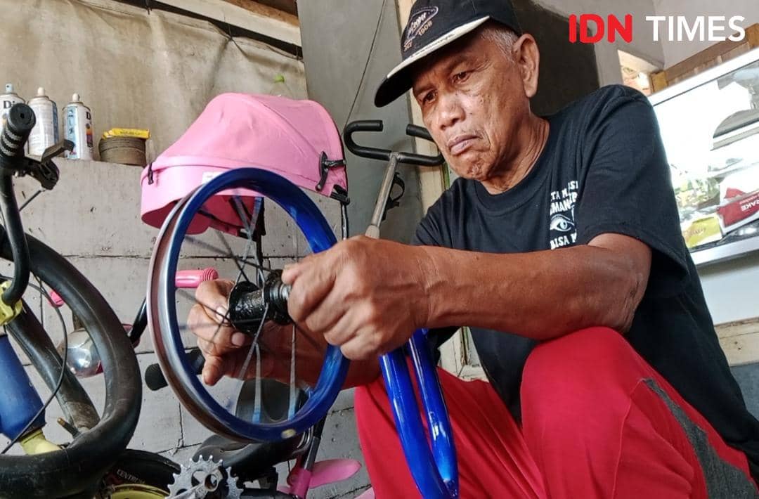 Seorang pria lanjut usia mengenakan topi hitam dan celana merah sedang memasang pelk sepeda berwarna biru di bengkel sederhana.