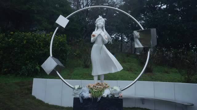 Patung Barbie Hsu berwarna putih berdiri di tengah lingkaran logam dengan elemen kubus, dikelilingi taman hijau dan bunga di bagian bawah.