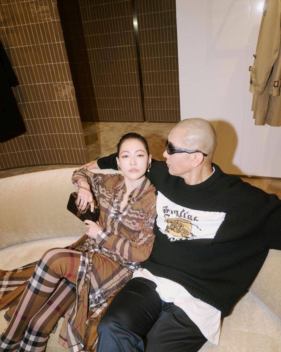 Seorang wanita berbusana motif kotak duduk di sofa bersama seorang pria berkacamata hitam mengenakan sweater hitam dan celana gelap.