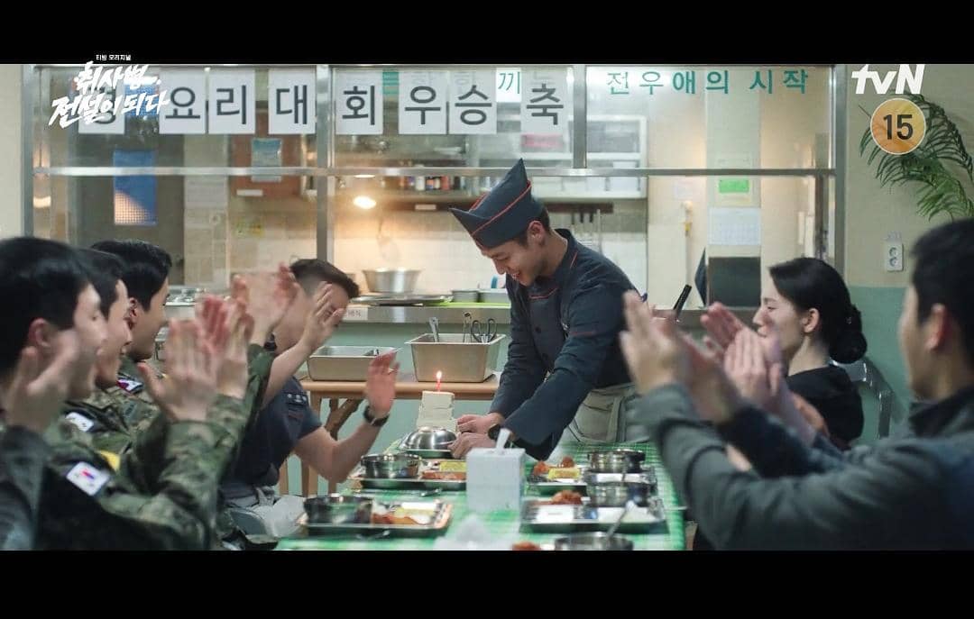 Seorang prajurit berpakaian koki menerima tepuk tangan dari rekan-rekannya di ruang makan militer dalam adegan drama The Legend of Kitchen Soldier.