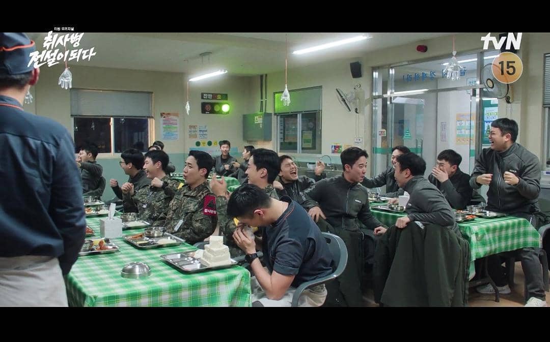 Para tentara sedang duduk di ruang makan barak dengan meja berlapis taplak hijau kotak, menikmati suasana santai dan bercanda bersama.