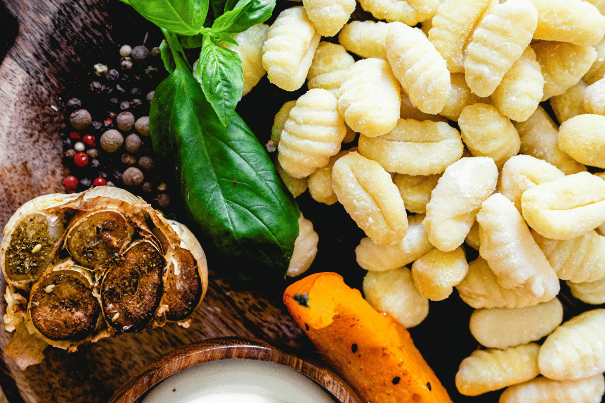 Tumpukan gnocchi mentah dengan bawang putih panggang, daun basil segar, lada hitam, dan potongan labu di atas talenan kayu.