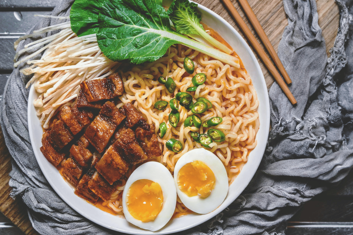 Semangkuk ramen dengan irisan daging perut babi panggang, telur rebus setengah matang, cabai hijau, jamur enoki, dan sayuran hijau.