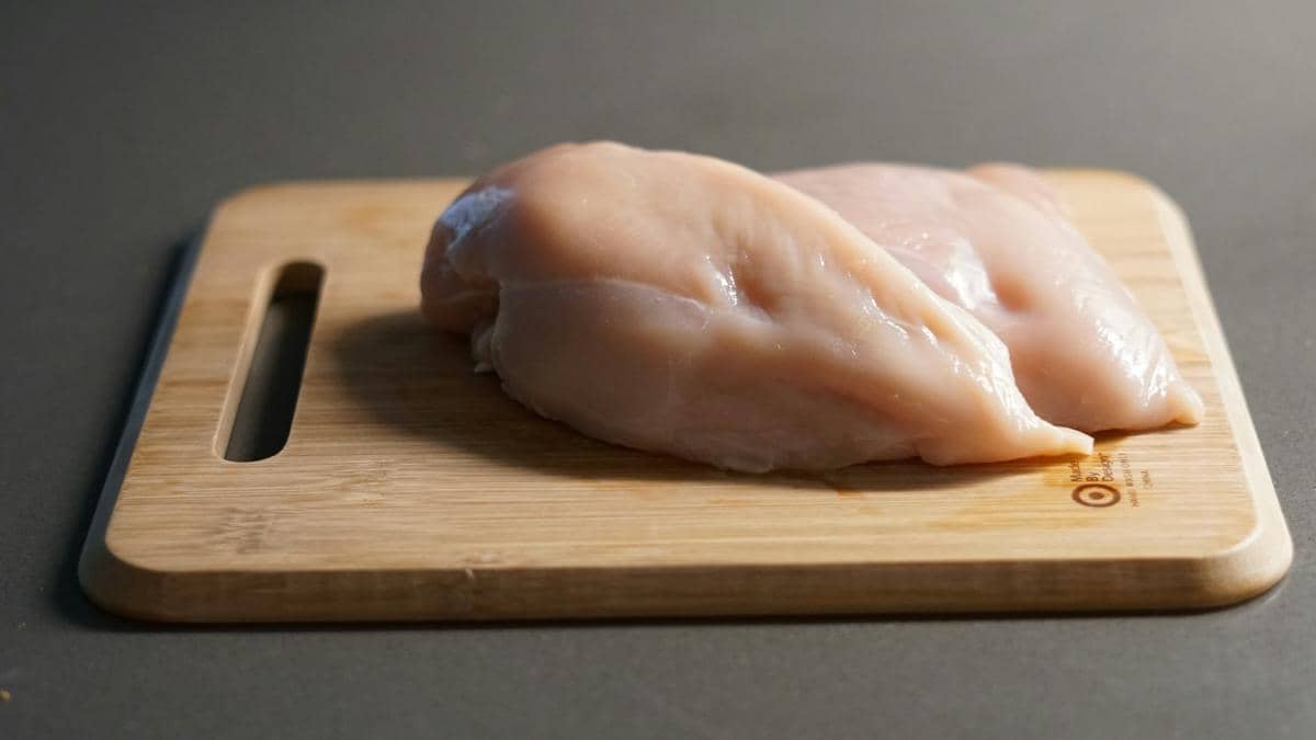 Dua potong daging ayam fillet mentah diletakkan di atas talenan kayu di permukaan meja dapur berwarna abu-abu.