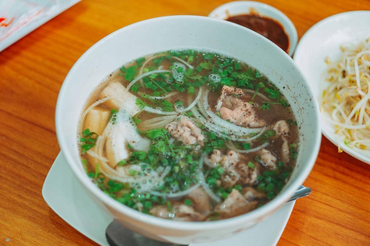 Semangkuk sukiyaki ala Jepang berisi irisan daging, sayuran, dan kuah hangat disajikan di atas meja kayu dengan pelengkap di sampingnya.