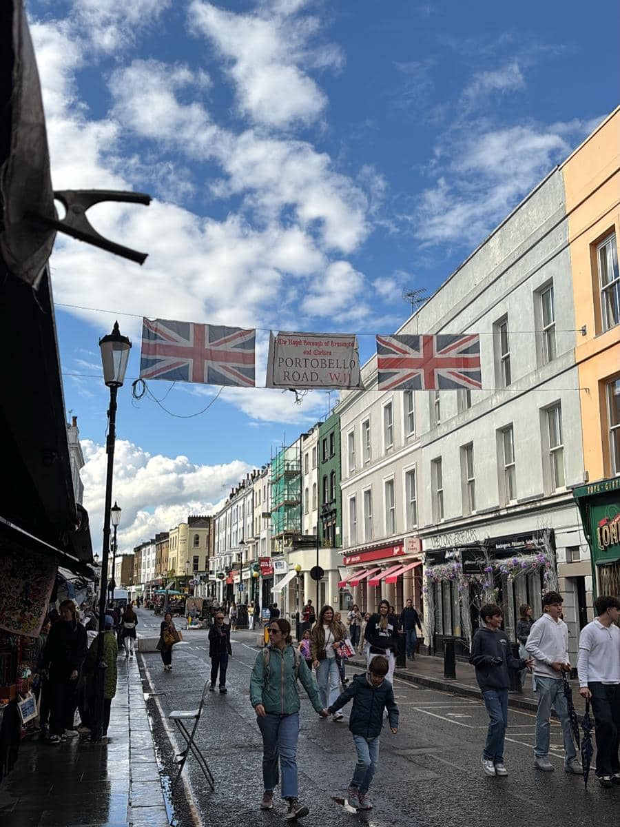 Suasana jalan Portobello Road di London dengan bendera Inggris tergantung di atas jalan dan orang-orang berjalan santai di bawah langit biru berawan.
