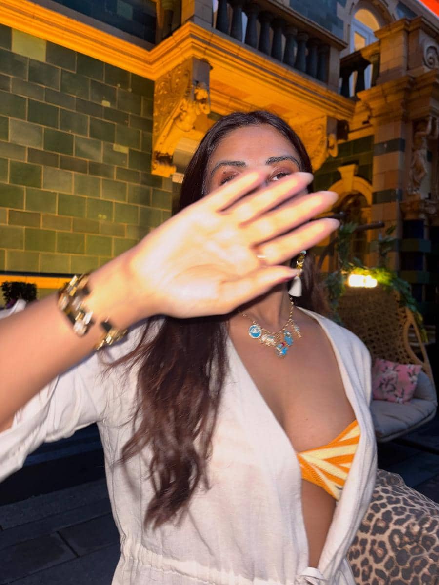 Seorang wanita berambut panjang mengenakan pakaian putih dan bikini oranye menutupi wajahnya dengan tangan di area luar ruangan bercahaya hangat.