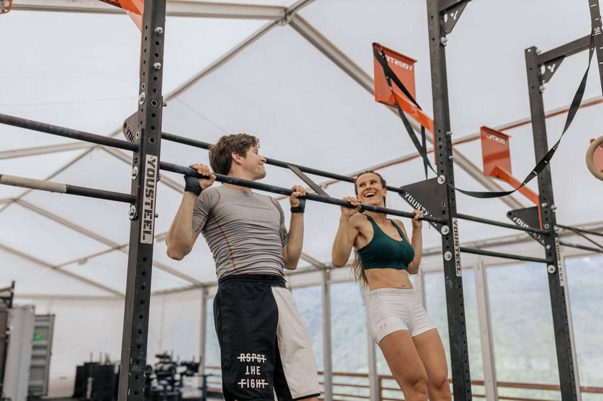Seorang pria dan wanita berolahraga bersama melakukan pull-up di gym dengan rangka besi di bawah tenda terbuka.