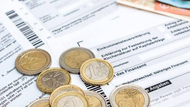 Sejumlah uang koin pecahan Euro berserakan di atas lembaran dokumen putih formulir deklarasi pajak berbahasa Jerman, memperlihatkan tulisan "Einkommensteuererklärung" dan kode batang hitam di sisi kiri atas.