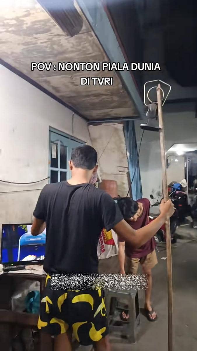 Dua pria berusaha memperbaiki posisi antena televisi di ruangan sederhana untuk menonton siaran Piala Dunia di TVRI.