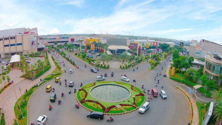 Bundaran besar di pusat Kota Karawang dengan taman hijau dan air mancur, dikelilingi kendaraan serta gedung pusat perbelanjaan modern.