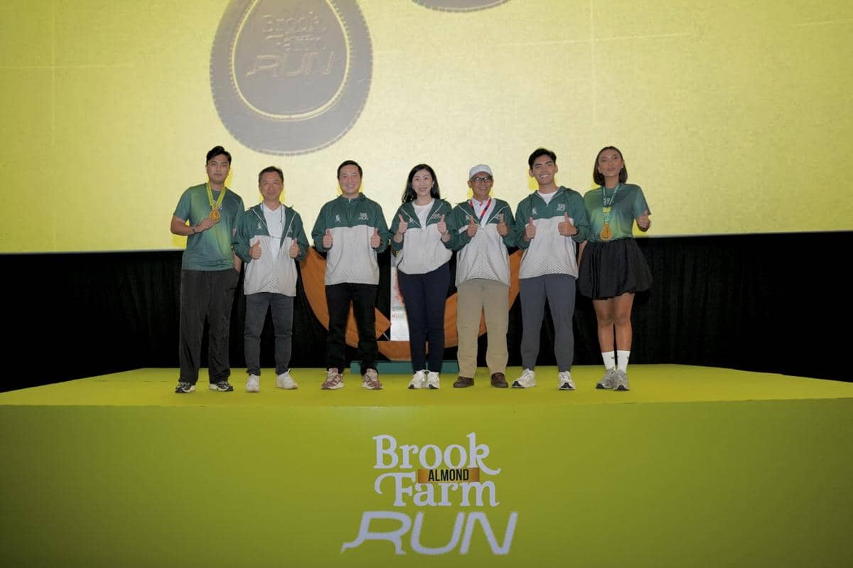 Tujuh orang mengenakan jaket hijau dan putih berpose di atas panggung dengan latar logo BrookFarm Almond Run 2026.