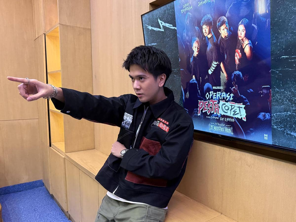 Seorang pria mengenakan jaket hitam berpose menunjuk ke depan di depan layar yang menampilkan poster film Operasi Pesta Copet.